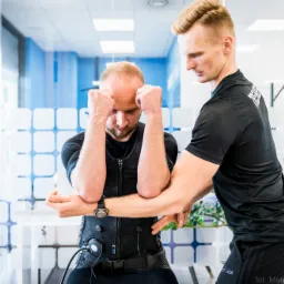 Trener personalny asystuje mężczyźnie w czarnym kombinezonie EMS podczas ćwiczeń, z widocznym zegarkiem na nadgarstku klienta, na tle biura z rośliną doniczkową i zegarem na ścianie.