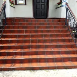 Schody zewnętrzne wyłożone brązową, błyszczącą terakotą z czarną, metalową balustradą i drewnianym poręczą, prowadzące do ciemnych drzwi wejściowych.