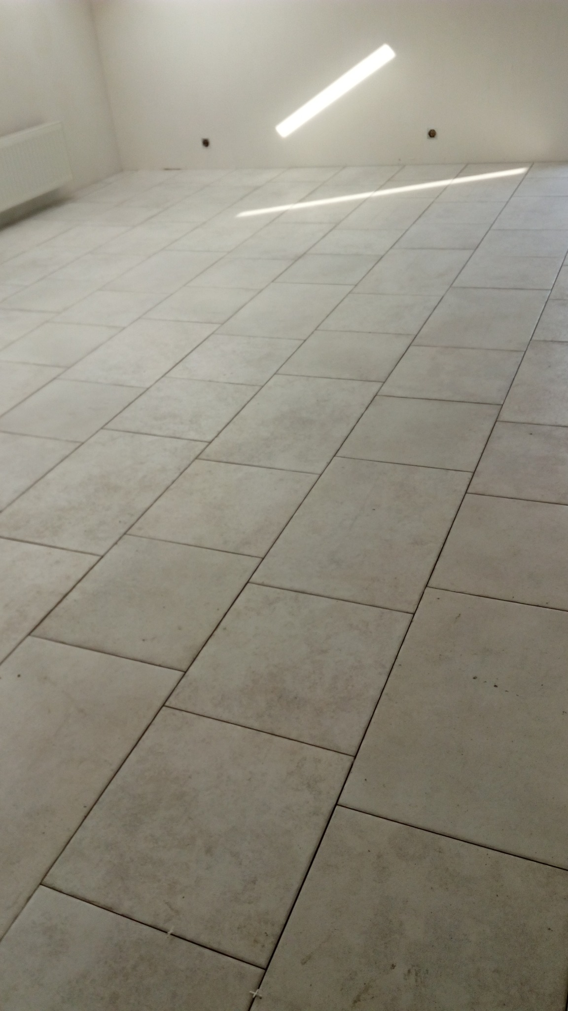 Jasne płytki podłogowe ułożone w regularny wzór, widoczne szczeliny między płytkami, na ścianie ślady po wierceniu, wpadające promienie słoneczne.