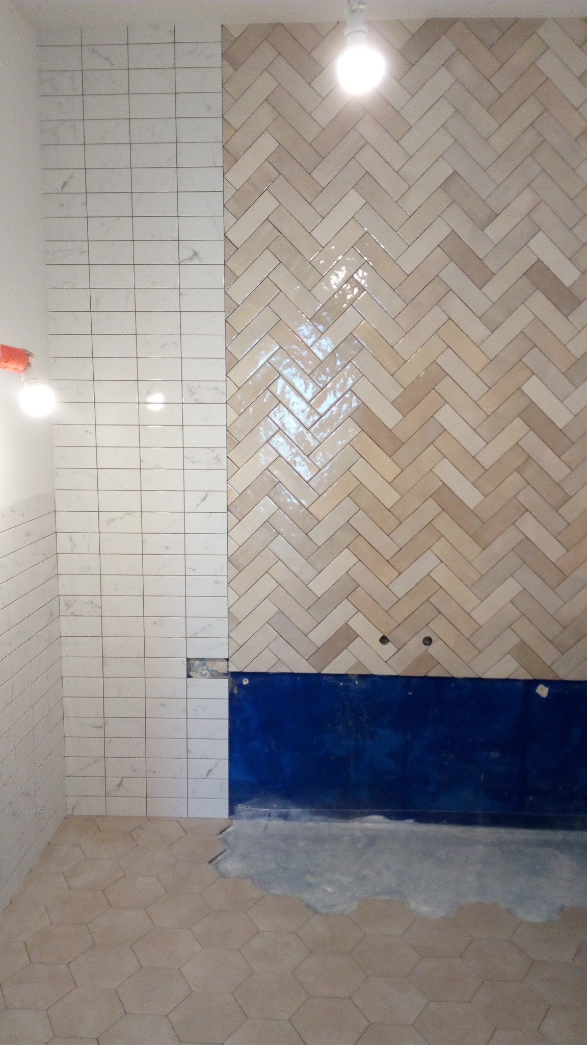 Ściana łazienki w trakcie układania płytek: część wykończona w białe prostokąty imitujące marmur, obok beżowa mozaika w jodełkę, poniżej niebieskie zabezpieczenie przed wilgocią, na podłodze...