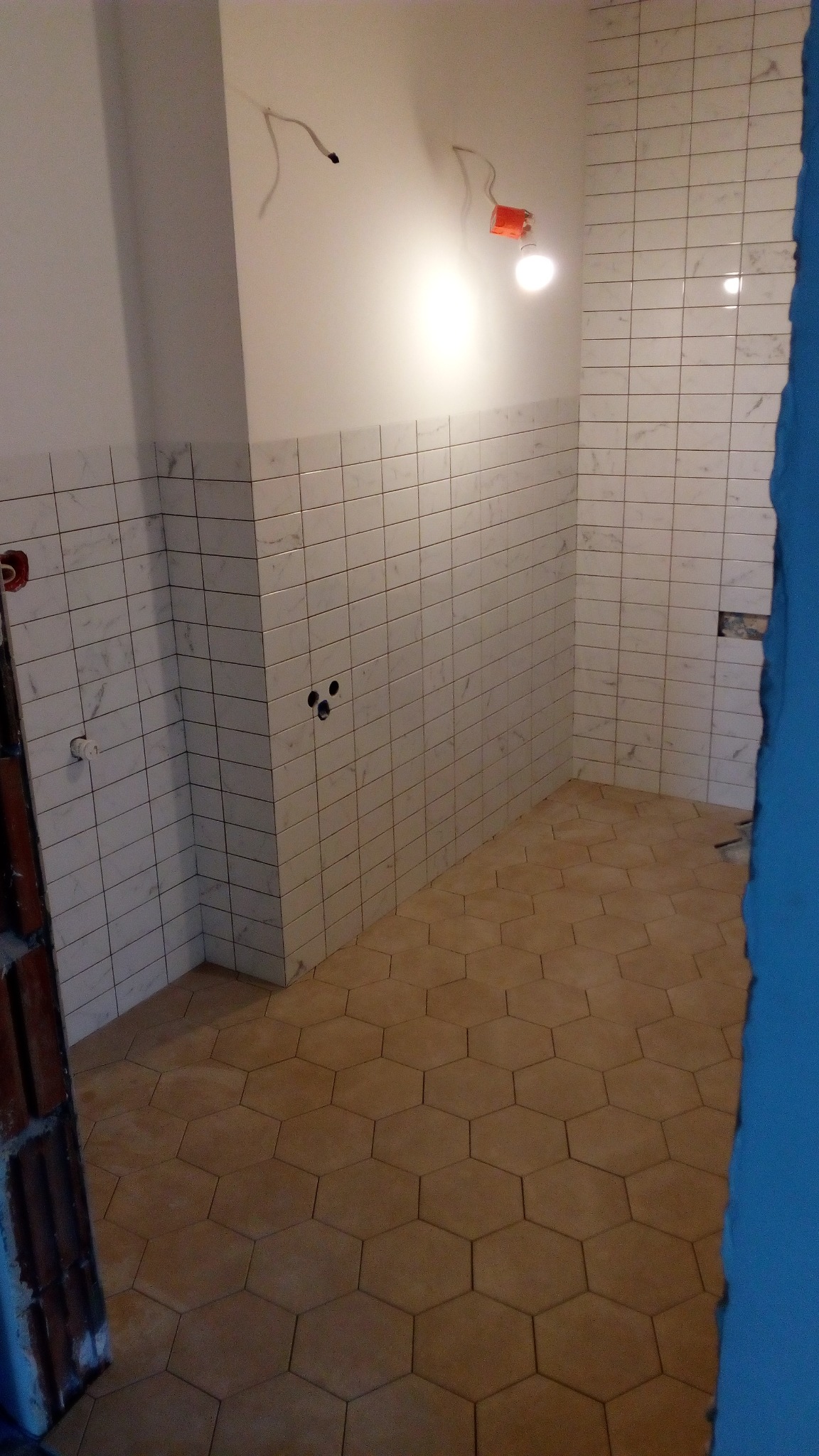 Wnętrze łazienki w trakcie prac wykończeniowych, widoczne płytki ceramiczne na ścianach do połowy wysokości i heksagonalne płytki na podłodze, niedokończone instalacje elektryczne na ścianie.