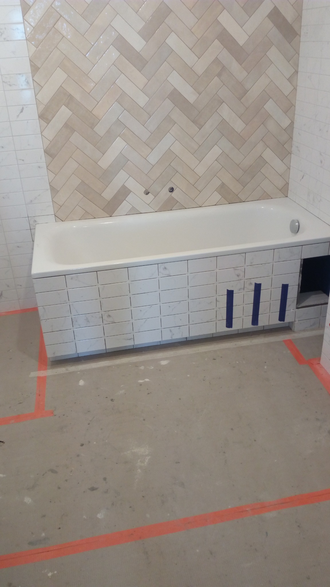 Obudowana płytkami wanna w trakcie prac, z widocznymi niebieskimi paskami taśmy mocującej i fragmentem ściany z wzorem jodełki z płytek w odcieniach beżu i szarości.