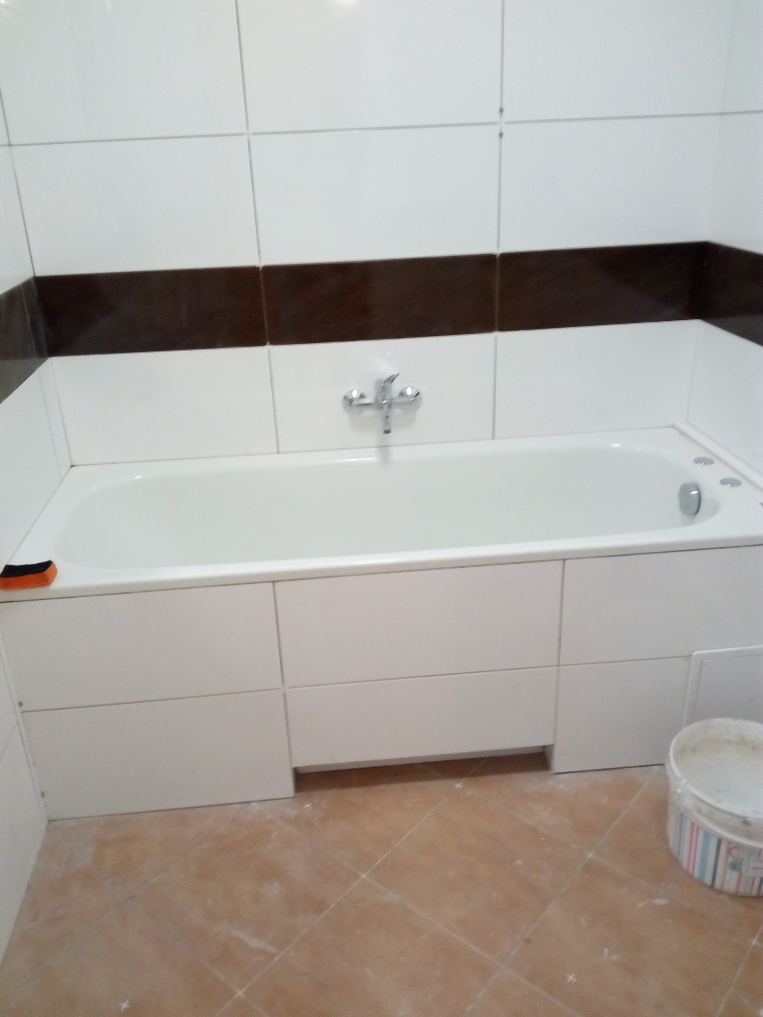 Biała wanna obudowana płytkami, z baterią chromowaną, na tle białych płytek z brązowym pasem, podłoga w terakocie, widoczne ślady po remoncie i wiadro po farbie.