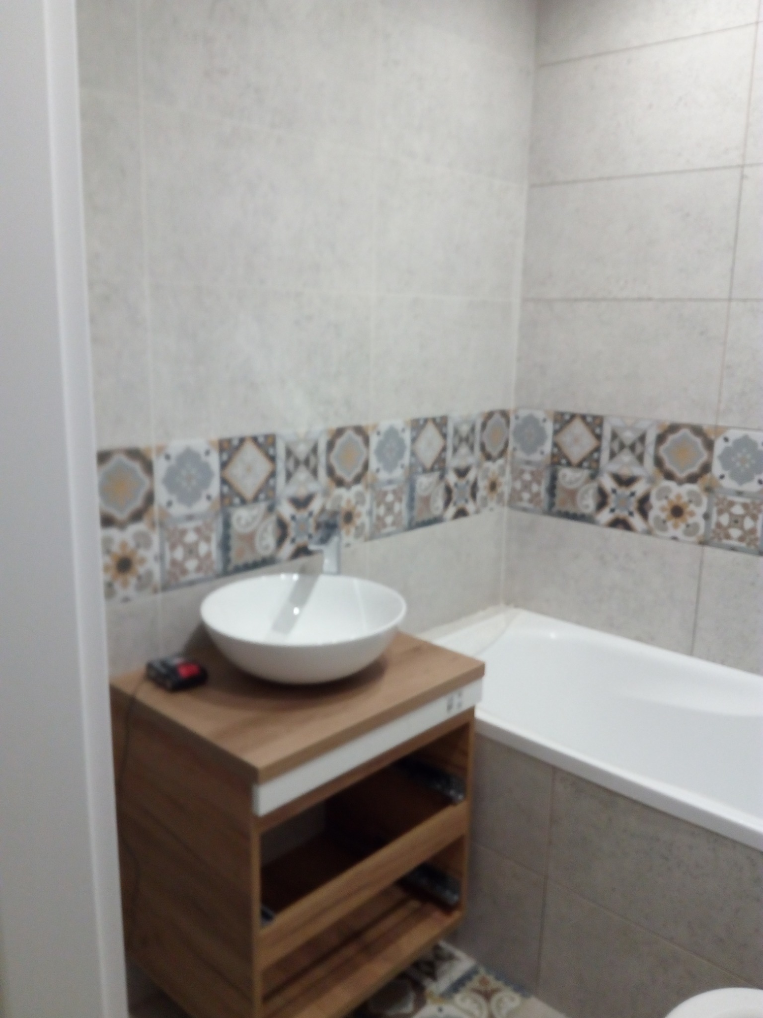 Wykończona łazienka z umywalką nablatową na drewnianej szafce z otwartymi półkami, ozdobny pas płytek na ścianie i biała wanna.