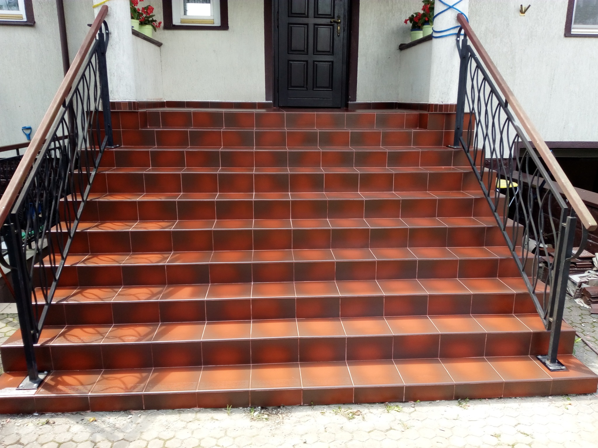 Schody zewnętrzne wyłożone brązową, błyszczącą terakotą z czarną, metalową balustradą i drewnianym poręczą, prowadzące do ciemnych drzwi wejściowych.