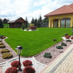 Zadbany trawnik przed domem z równo przyciętą trawą, obrzeżami z kamieni i niskimi krzewami ozdobnymi, oświetlony lampami ogrodowymi wzdłuż ścieżki z kostki brukowej.