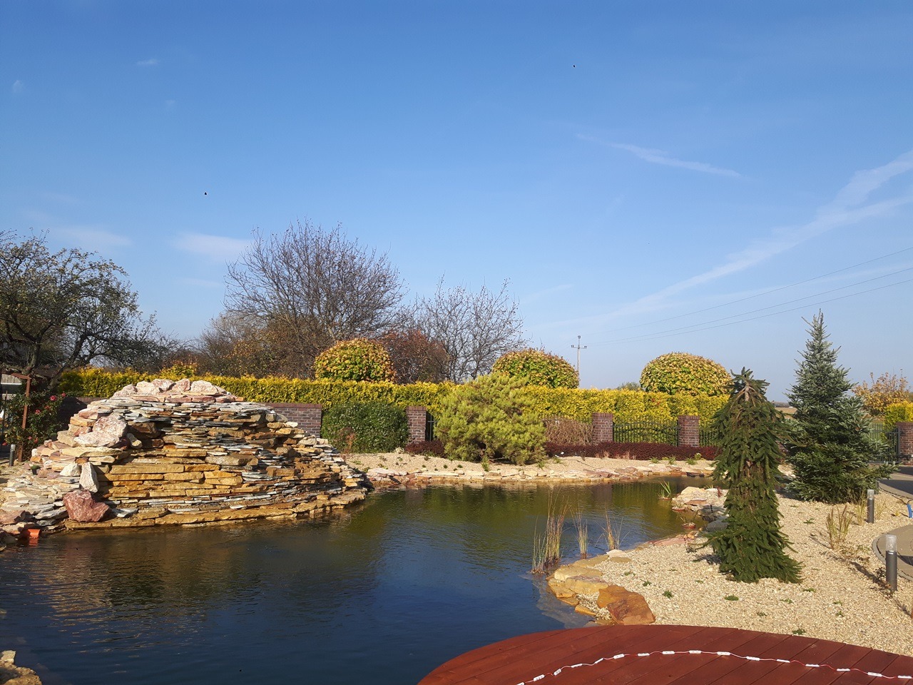 Oczko wodne z kaskadą z kamieni warstwowych, otoczone ozdobnym żwirem i roślinnością, w tle żywopłot i niebieskie niebo.