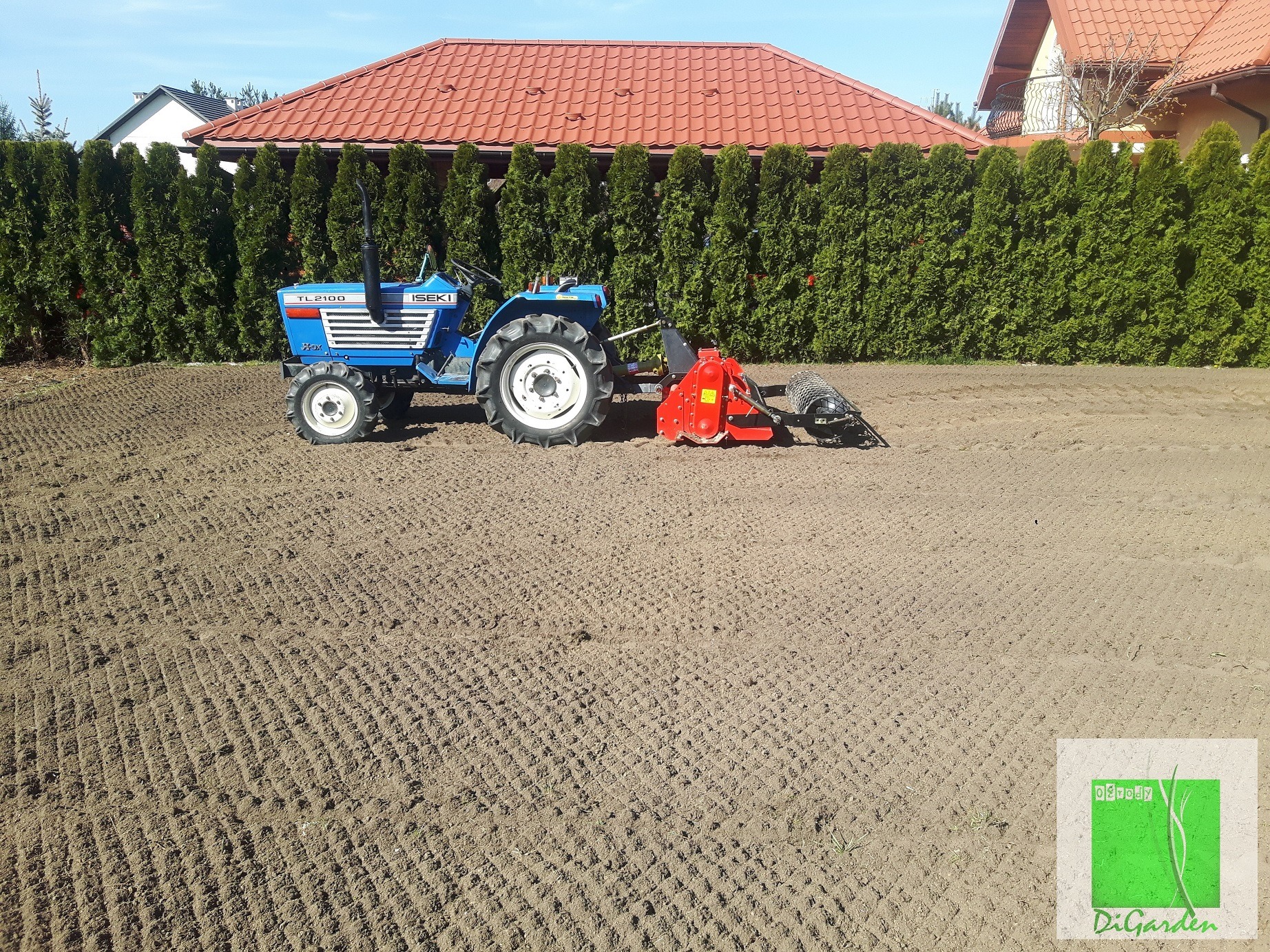 Niebieski traktor Iseki TL2100 z czerwonym glebogryzem na przygotowanym do siewu trawniku, rząd tuj w tle, logo firmy w prawym dolnym rogu.