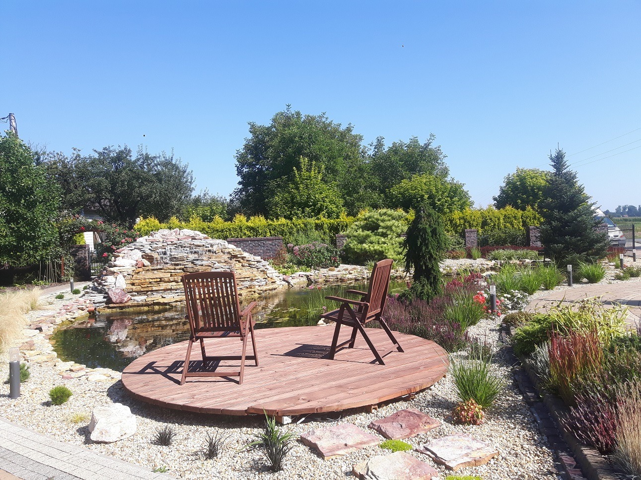 Oczko wodne z kaskadą i drewnianym pomostem z dwoma krzesłami, otoczone różnorodną roślinnością, w słoneczny dzień.