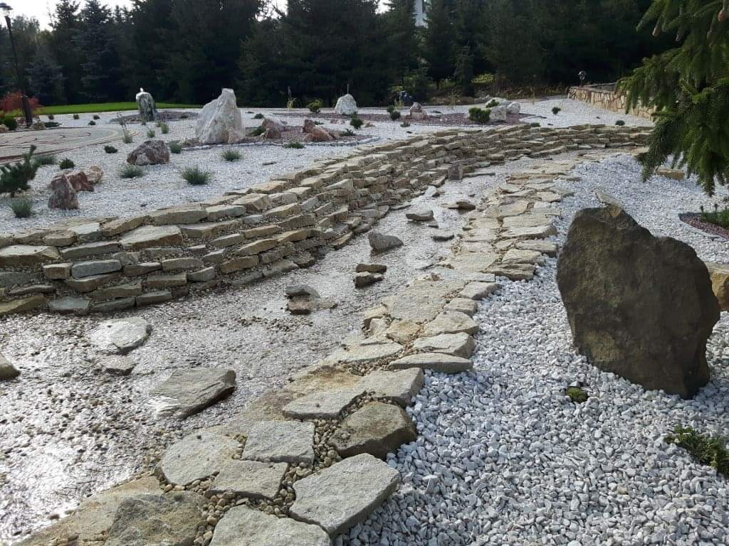 Aranżacja ogrodu z kamienną rzeczką, białym żwirem i dekoracyjnymi głazami, widok z poziomu ziemi.