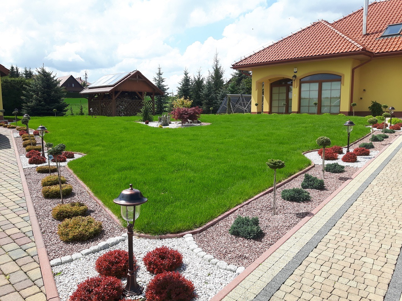 Zadbany trawnik przed domem z równo przyciętą trawą, obrzeżami z kamieni i niskimi krzewami ozdobnymi, oświetlony lampami ogrodowymi wzdłuż ścieżki z kostki brukowej.