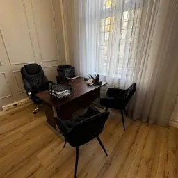 Nowoczesne biuro: biurko z dokumentami, fotel biurowy i dwa krzesła dla klientów, jasne drewniane podłogi, okno z białymi zasłonami, stonowana kolorystyka.