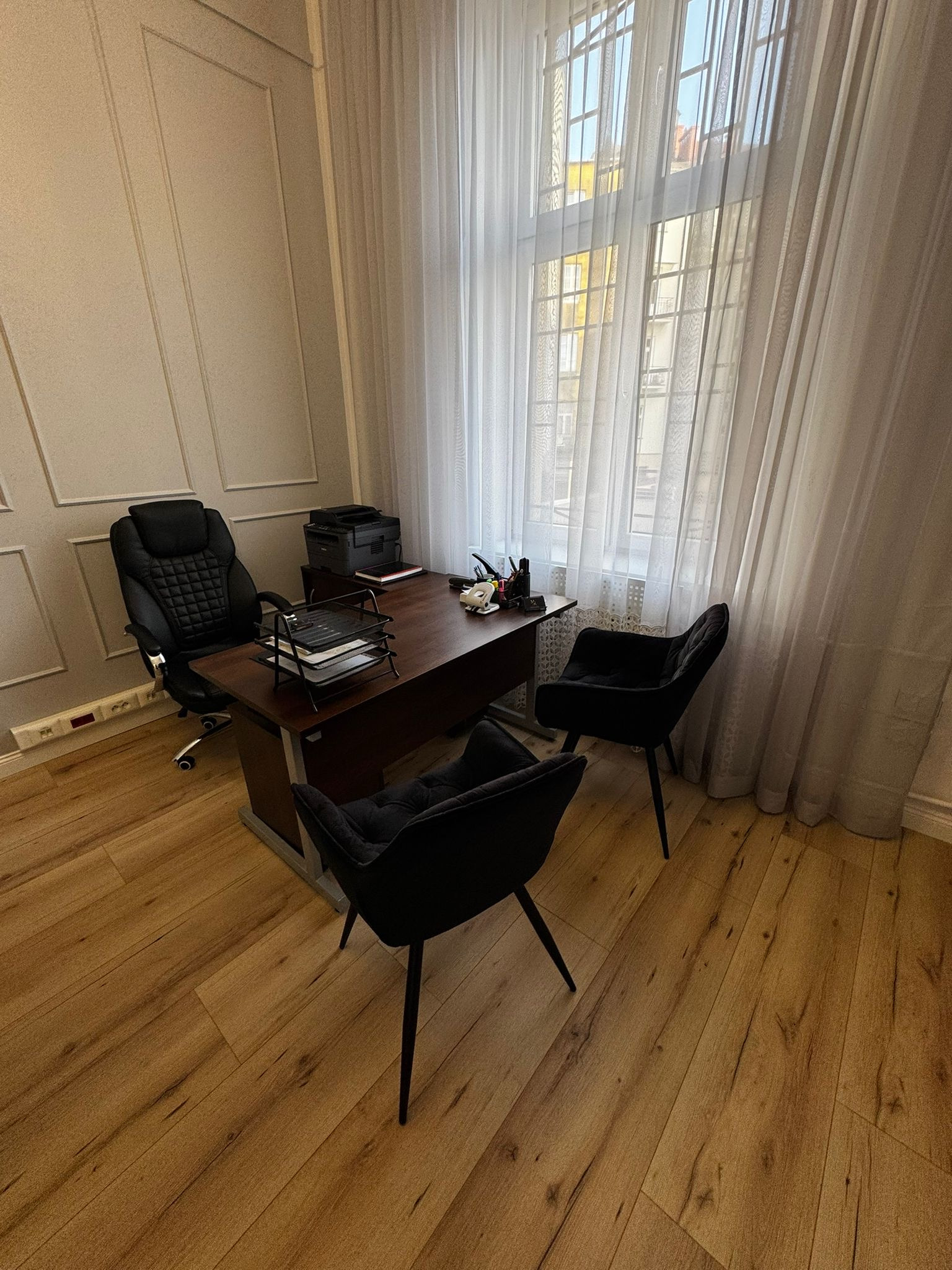 Nowoczesne biuro: biurko z dokumentami, fotel biurowy i dwa krzesła dla klientów, jasne drewniane podłogi, okno z białymi zasłonami, stonowana kolorystyka.