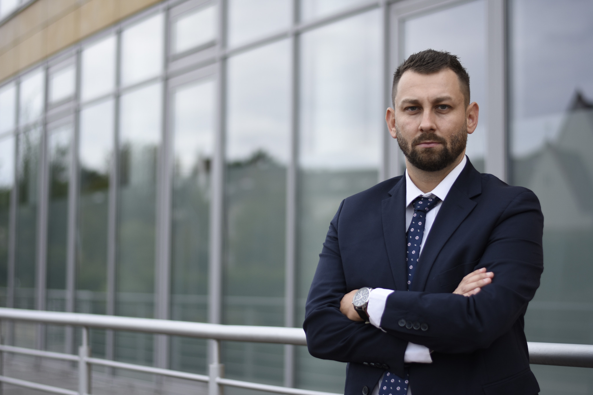 Portret mężczyzny w garniturze, z brodą, stojącego na tle nowoczesnego budynku z przeszkloną fasadą, z założonymi rękami i zegarkiem na nadgarstku.