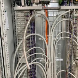 Otwarta szafa sterownicza z licznymi przewodami, modułami i elementami łączeniowymi; widoczne przewody w kolorach białym, pomarańczowym i srebrnym, oraz moduły Siemens.