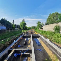 Wykop pod fundamenty domu jednorodzinnego z widocznym zbrojeniem, izolacją pionową i częściowo ułożonymi rurami instalacyjnymi, w tle zabudowa jednorodzinna i błękitne niebo.