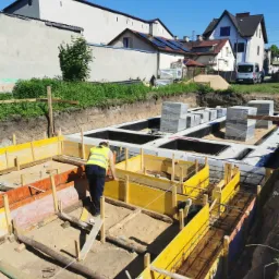 Pracownik w żółtej kamizelce odblaskowej sprawdza szalunki fundamentów domu jednorodzinnego, widoczne zbrojenia i ułożone bloczki fundamentowe, w tle zabudowa jednorodzinna.