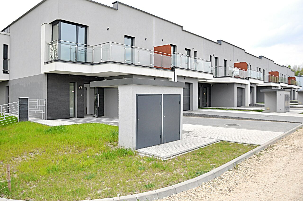 Szereg nowoczesnych, dwukondygnacyjnych domów z balkonami, wykończonych w szarości, z elementami dekoracyjnymi w kolorze pomarańczowym na balkonach, widoczne skrzynki na media przed każdym domem.