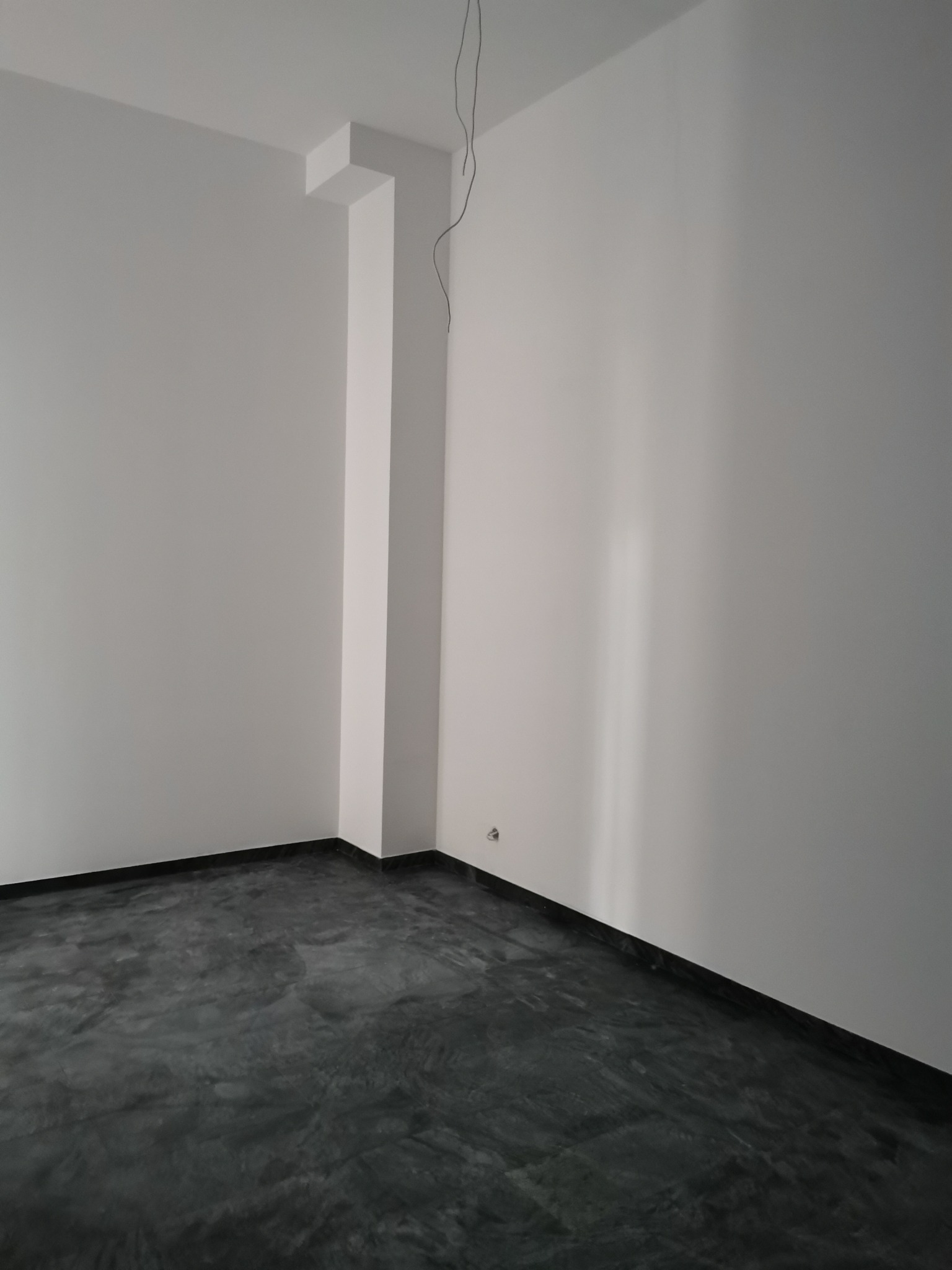 Pusty pokój z szarym, marmurkowym betonem na podłodze, świeżo pomalowane białe ściany i wystający filar z kablem zwisającym z sufitu.