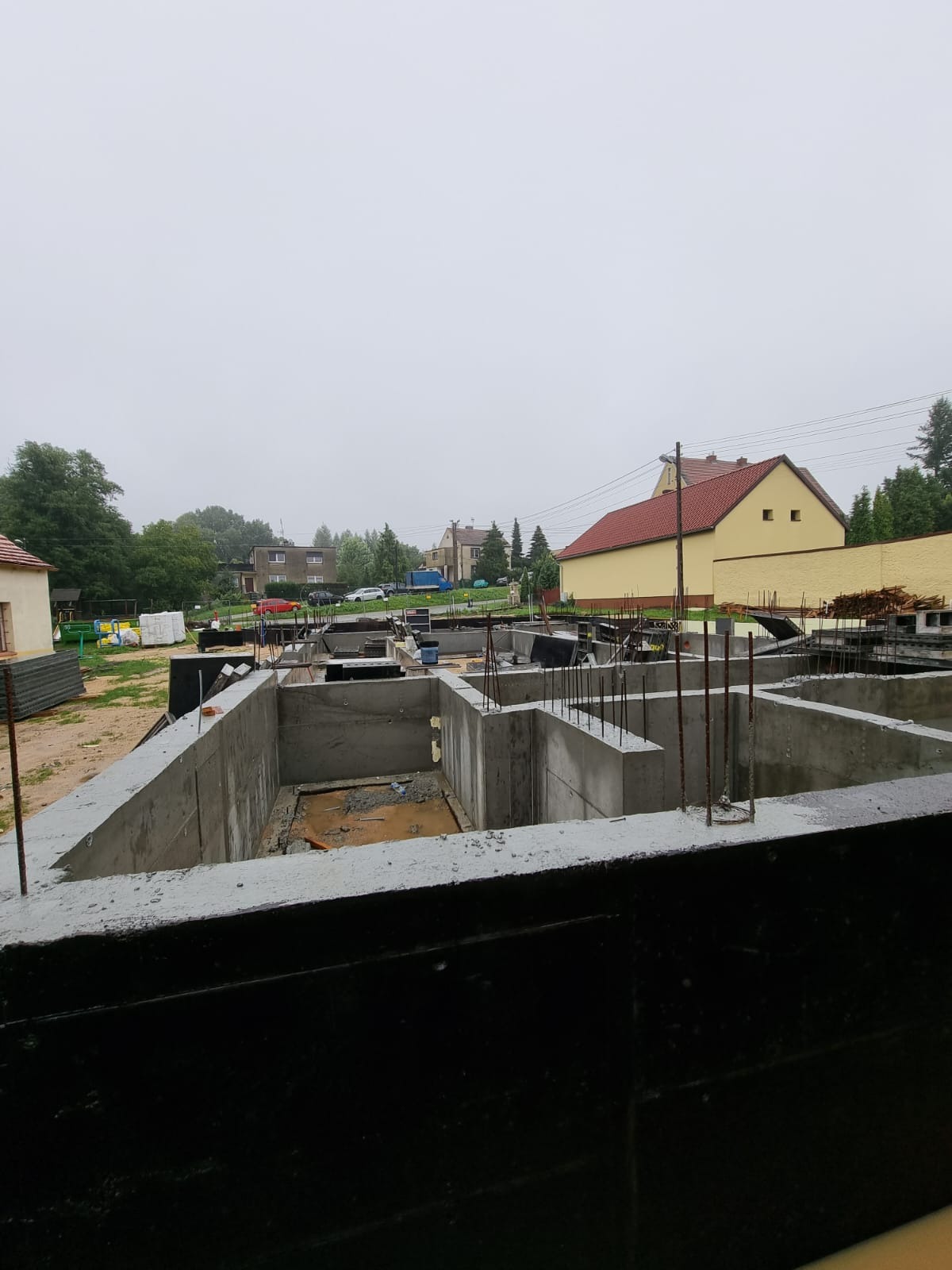 Betonowy fundament domu jednorodzinnego z wystającymi prętami zbrojeniowymi, widok z góry na tle zabudowy mieszkalnej w pochmurny dzień.