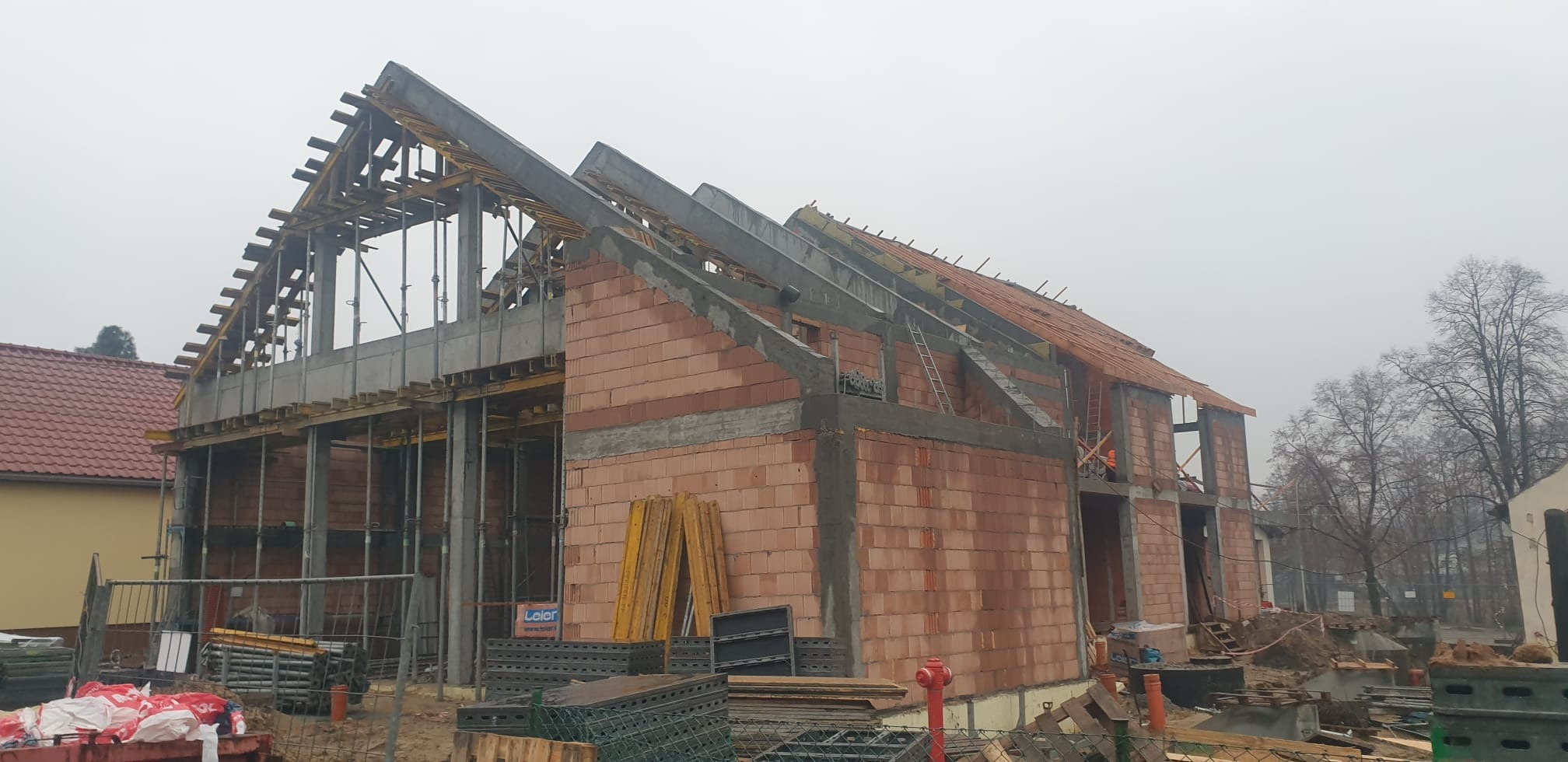 Budowa domu murowanego w trakcie realizacji: ściany z czerwonej cegły, częściowo wylane betonowe elementy konstrukcyjne, drewniane szalunki dachu, widoczne rusztowania i materiały budowlane...