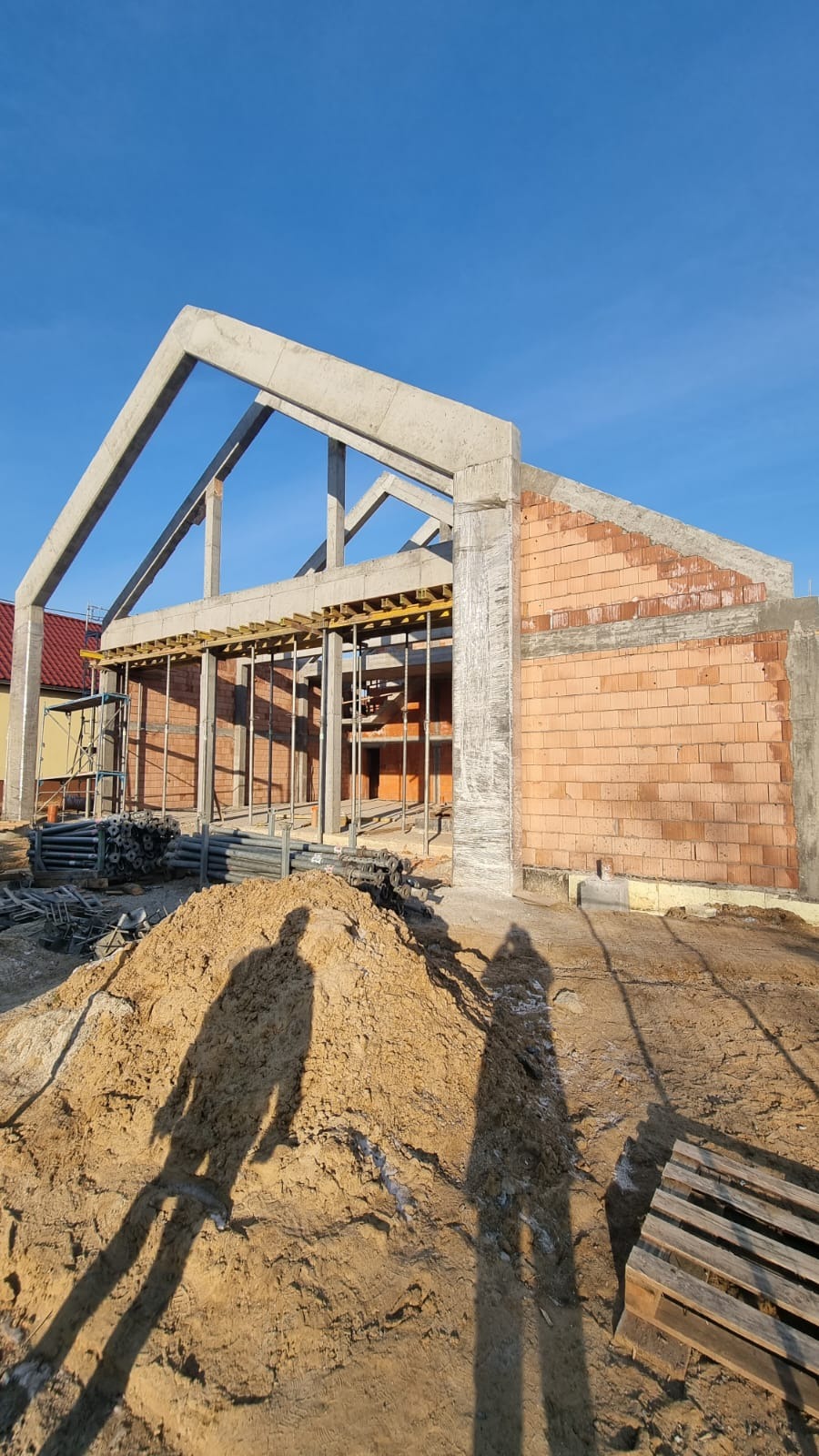 Surowy stan budowy domu z betonu i cegły, widoczne szalunki i zbrojenia, na pierwszym planie hałda piasku z cieniami dwóch osób.