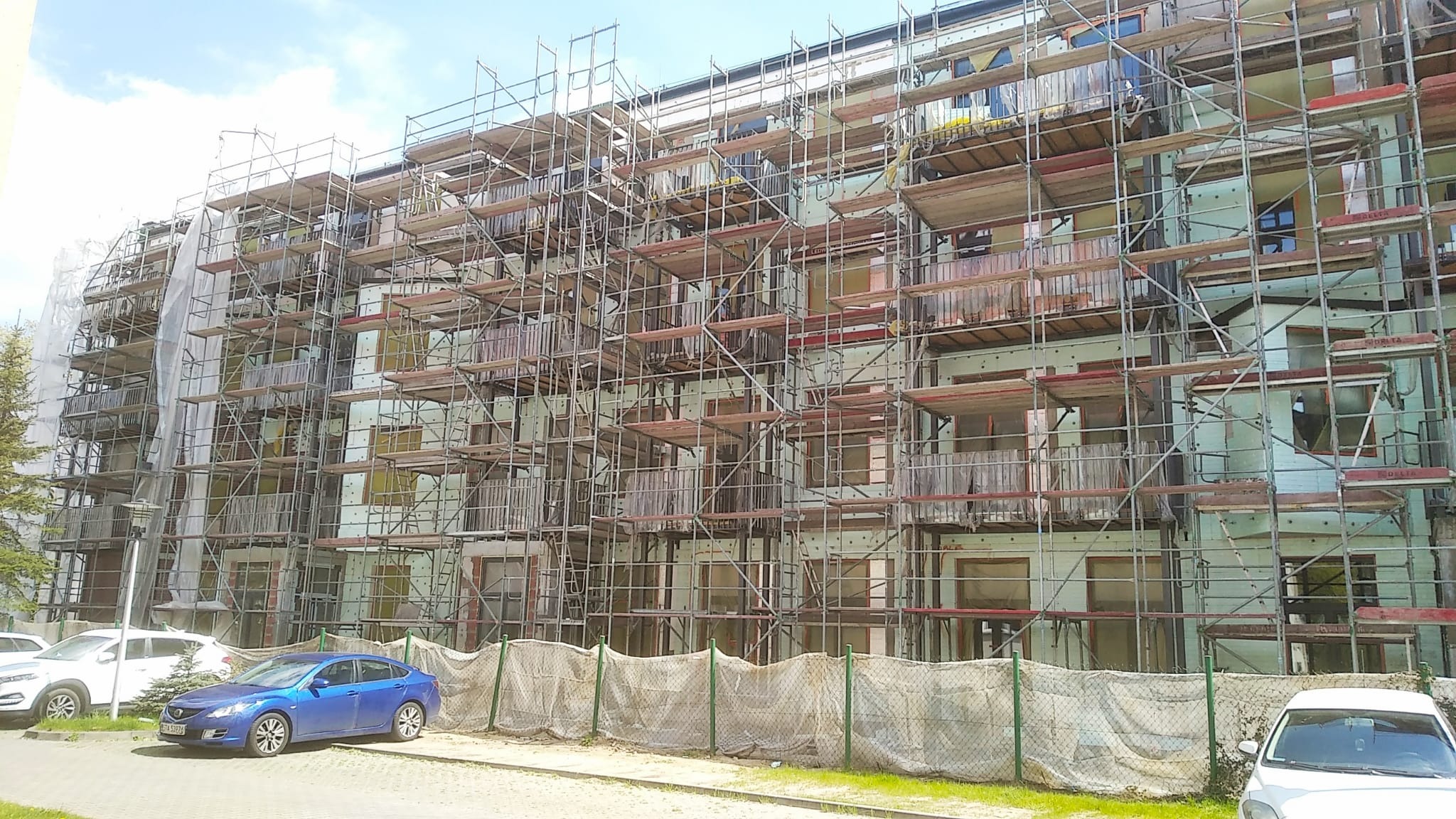 Wielopiętrowy budynek mieszkalny w trakcie ocieplania, pokryty rusztowaniami i częściowo zabezpieczony siatką, widoczny zaparkowany niebieski samochód.