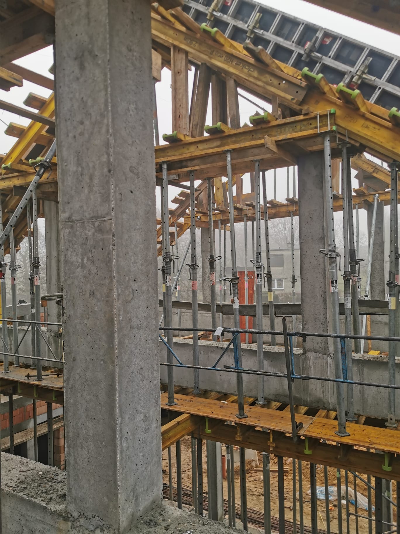 Wznoszenie konstrukcji żelbetowej budynku: szalunki drewniane podtrzymywane metalowymi stemplami, widoczne zbrojenie i betonowe słupy w surowym stanie, w tle zarys zabudowań w lekkiej mgle.