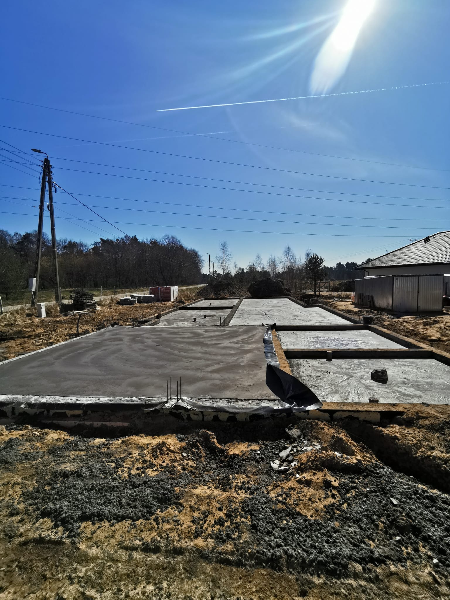 Wylany fundament z betonu na budowie domu jednorodzinnego, widoczne wystające pręty zbrojeniowe, w tle błękitne niebo z smugami kondensacyjnymi samolotu.