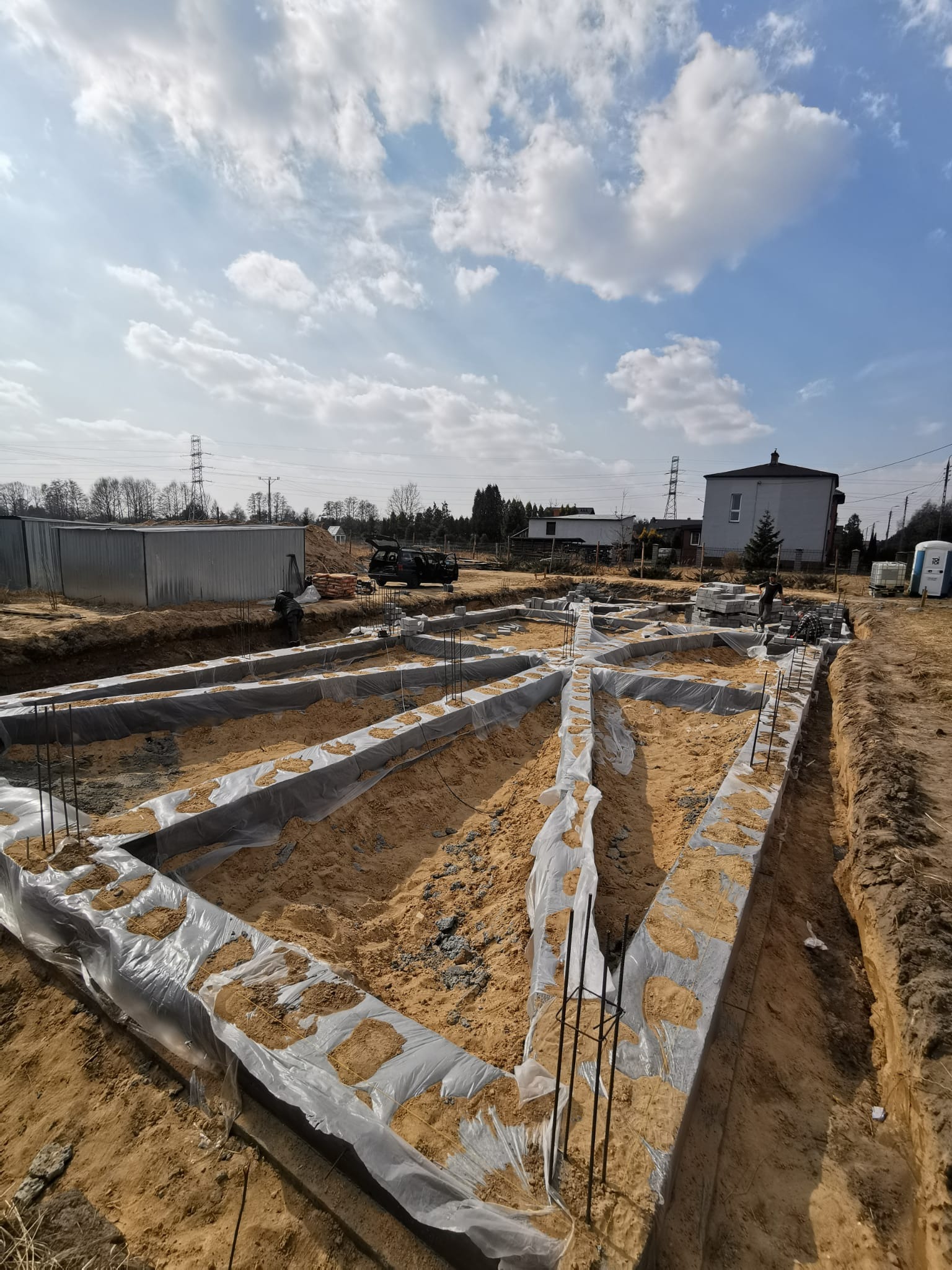 Zbrojone betonowe ławy fundamentowe pod budowę domu jednorodzinnego, zabezpieczone folią, z wystającymi prętami zbrojeniowymi, na piaszczystym podłożu, widok z góry.