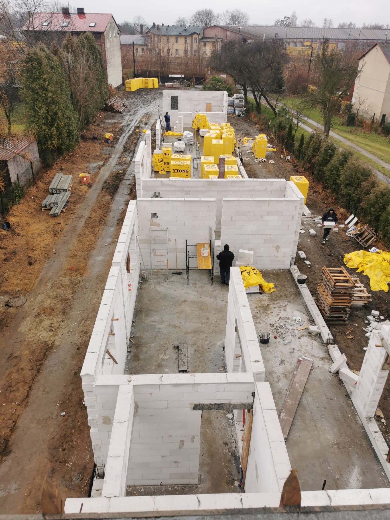 Budowa domu z białych bloczków Ytong, widok z góry na postęp prac, widoczne materiały budowlane w żółtych opakowaniach, drabina i pracownicy na placu budowy.