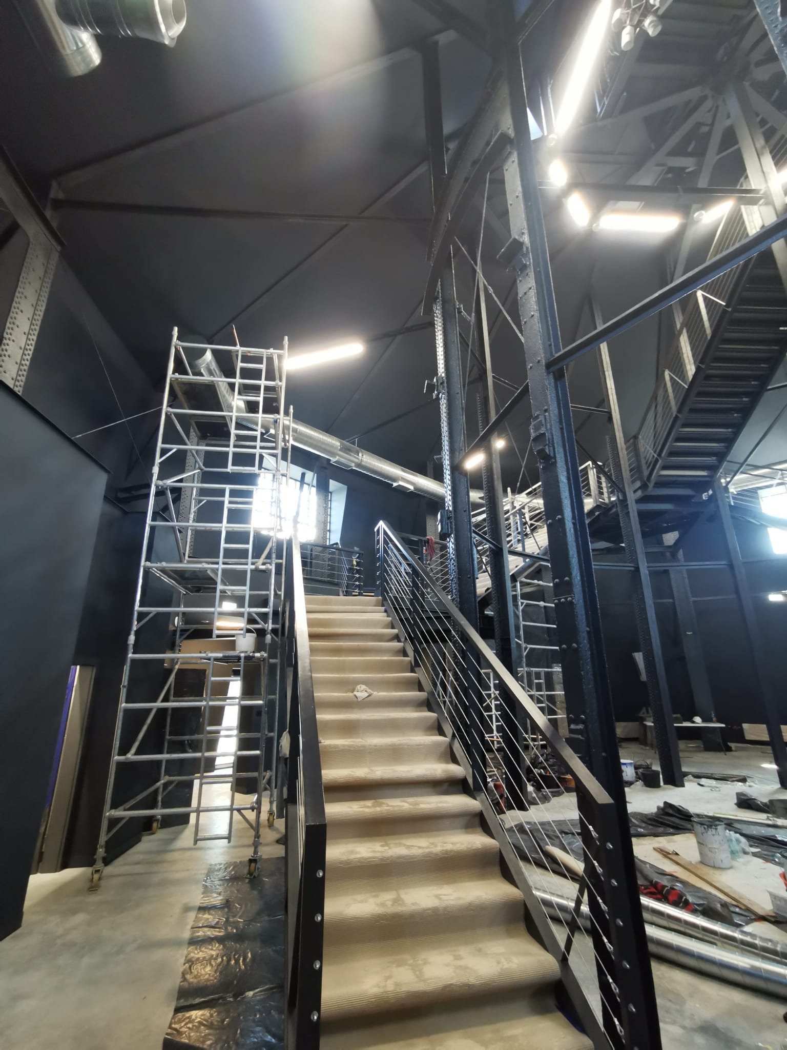 Wnętrze budynku w trakcie wykańczania: schody z metalową balustradą, rusztowanie, widoczne elementy konstrukcji stalowej i instalacji wentylacyjnej, czarne ściany i sufit.
