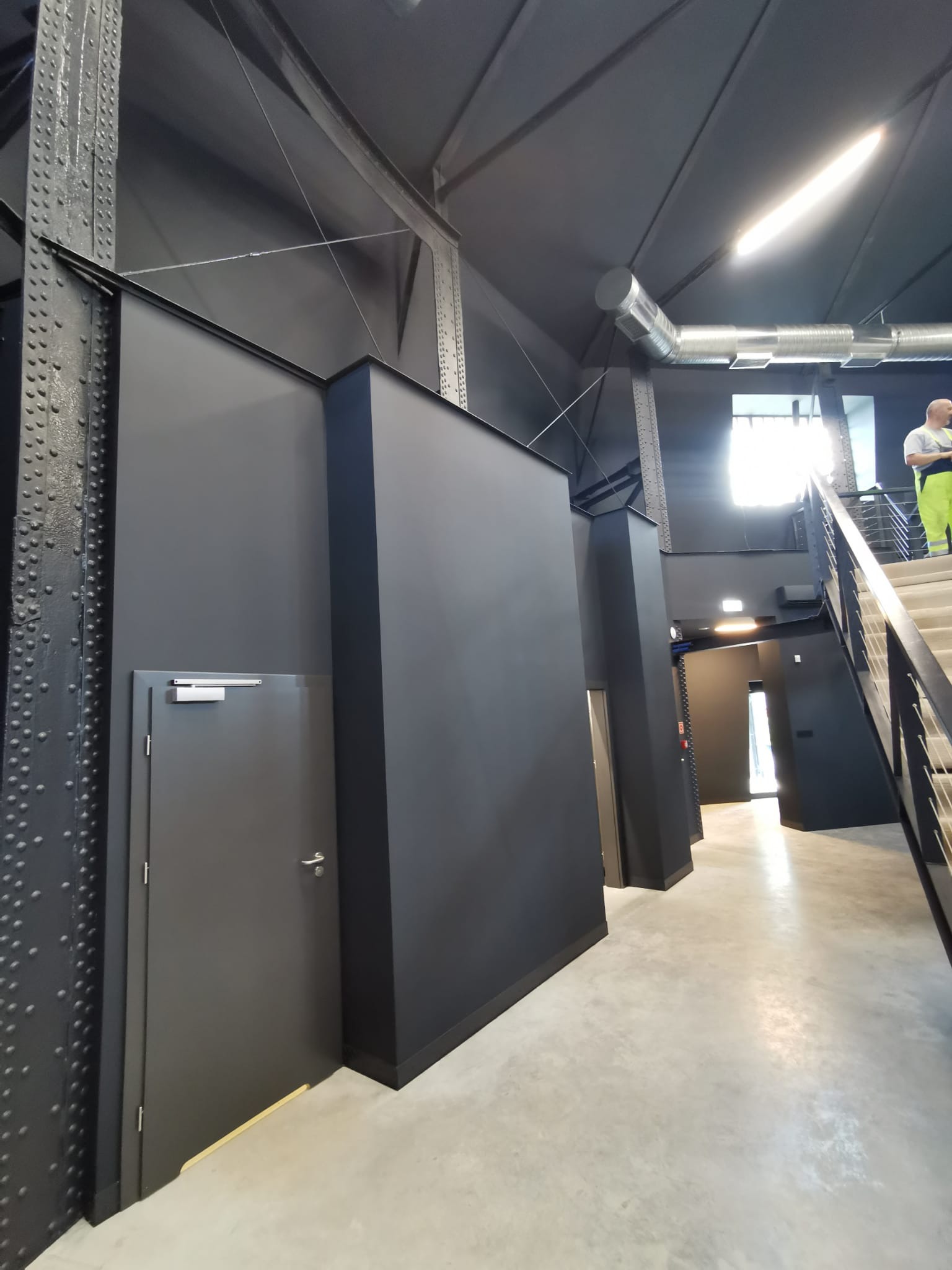 Wnętrze budynku w stylu industrialnym z czarnymi ścianami, metalowymi belkami konstrukcyjnymi z nitami, betonową posadzką i schodami z metalową balustradą; widoczny fragment instalacji...
