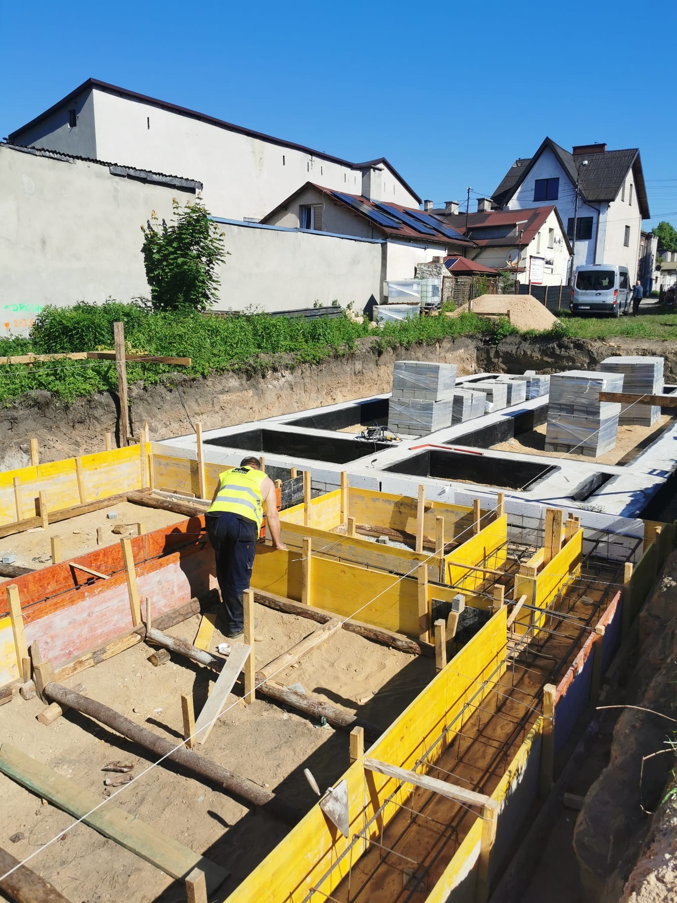 Pracownik w żółtej kamizelce odblaskowej sprawdza szalunki fundamentów domu jednorodzinnego, widoczne zbrojenia i ułożone bloczki fundamentowe, w tle zabudowa jednorodzinna.