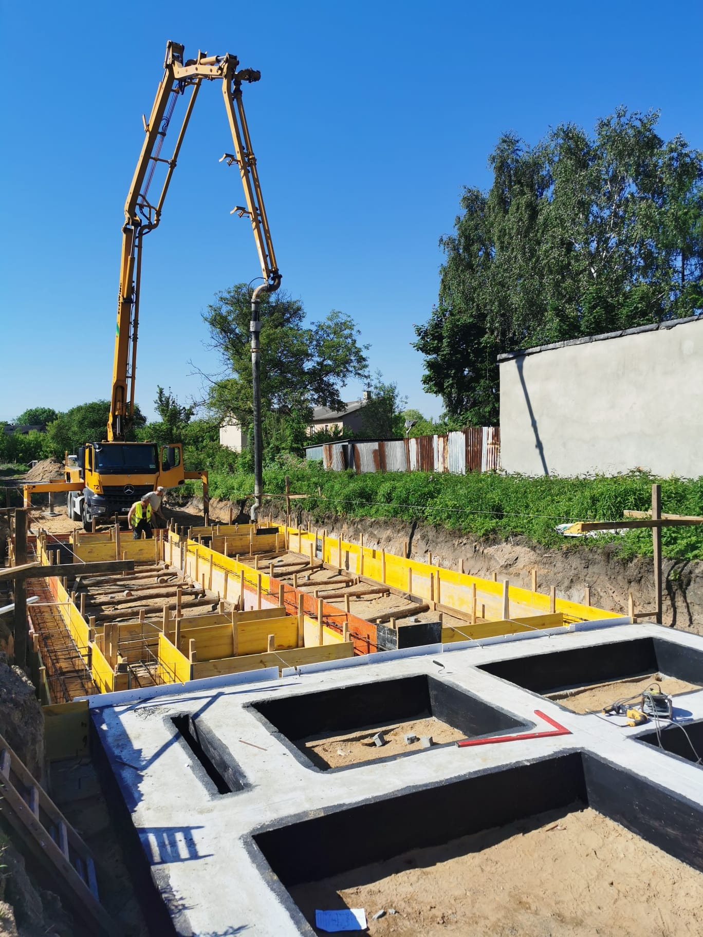 Wylewanie betonu do szalunków fundamentowych z użyciem pompy do betonu; widoczne zbrojenia, poziomica, oraz pracownicy w odblaskowych kamizelkach.