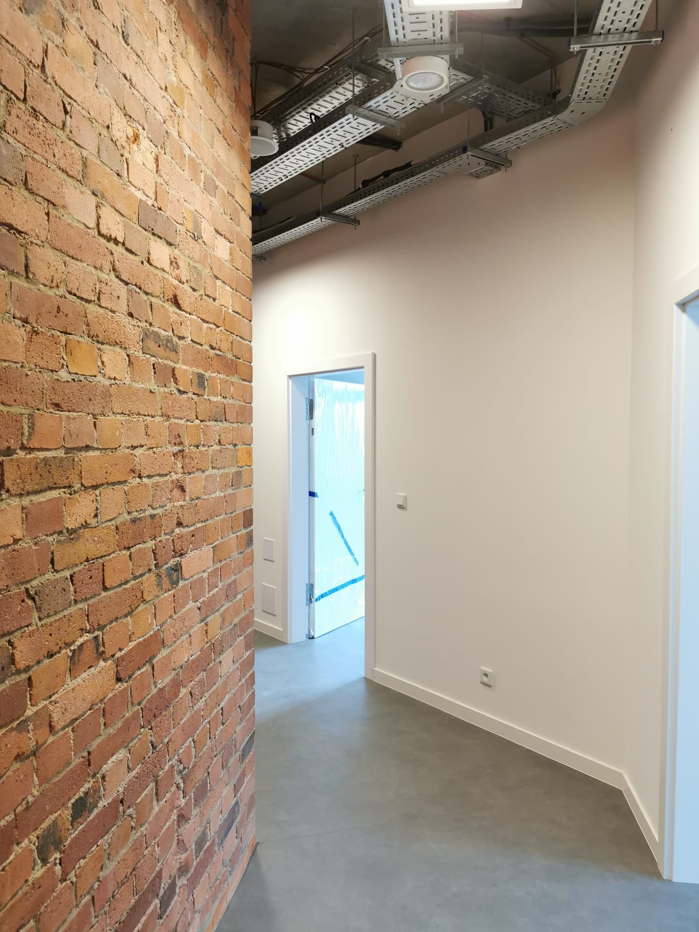 Korytarz w trakcie wykańczania: surowa ceglana ściana kontrastuje z gładką, pomalowaną na biało ścianą, widoczne koryta kablowe pod sufitem i drzwi zabezpieczone folią ochronną.