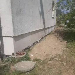 Świeżo otynkowana podmurówka budynku z widocznymi elementami izolacji przeciwwilgociowej, obok studzienka kanalizacyjna z betonową pokrywą i rurą spustową.