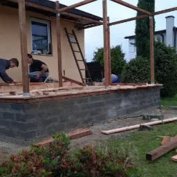 Budowa drewnianej konstrukcji zadaszenia tarasu: widoczne słupy, belki, częściowo ułożona podłoga na fundamencie z bloczków betonowych, drabina oparta o ścianę budynku, dwóch pracowników w trakcie...