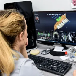 Kobieta z blond włosami w kucyku analizuje na monitorze trójwymiarowy model szczęki z zaznaczonymi implantami, widoczne narzędzia i materiały dentystyczne na biurku.