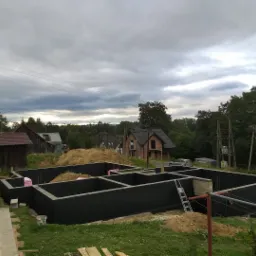 Wylane fundamenty domu jednorodzinnego z izolacją pionową, widoczny zarys ścian nośnych, częściowo zasypane piaskiem, drabina oparta o ścianę fundamentową, w tle zabudowa jednorodzinna i pochmurne...