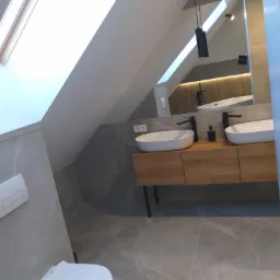 Nowoczesna łazienka na poddaszu ze skośnym oknem dachowym, umywalkami nablatowymi na drewnianej szafce, szarą marmurkową podłogą i ścianami oraz czarnymi detalami armatury.