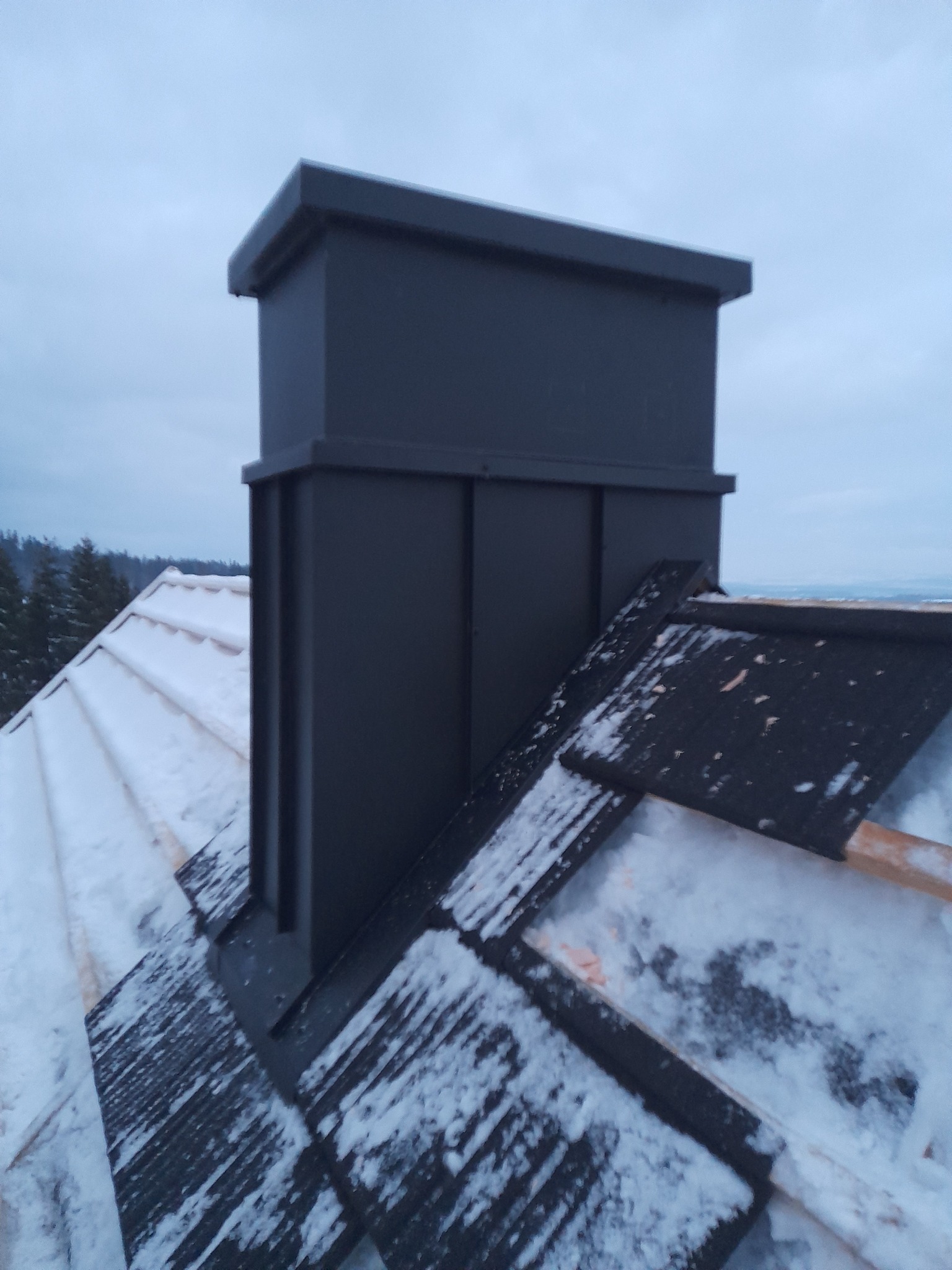 Nowoczesny, grafitowy komin systemowy na dachu pokrytym śniegiem, widok z góry w pochmurny dzień.