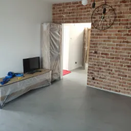 Szara, jednolita posadzka w nowym mieszkaniu z ceglaną ścianą, częściowo umeblowanym i zabezpieczonym meblami folią ochronną.