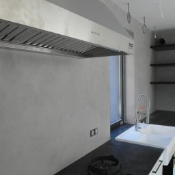 BLIKER sp. z o.o. - Kuchnia z wykończeniem ścian z mikrocementu w odcieniach szarości. Widoczny okap, zlew i czarne półki. Minimalistyczny design z nowoczesnym oświetleniem.