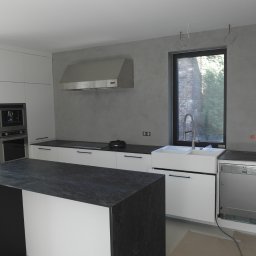 BLIKER sp. z o.o. - Nowoczesna kuchnia w trakcie wykończenia: białe szafki, czarny blat wyspy, ściana pokryta szarym mikrocementem. Widoczne niedokończone detale instalacji.