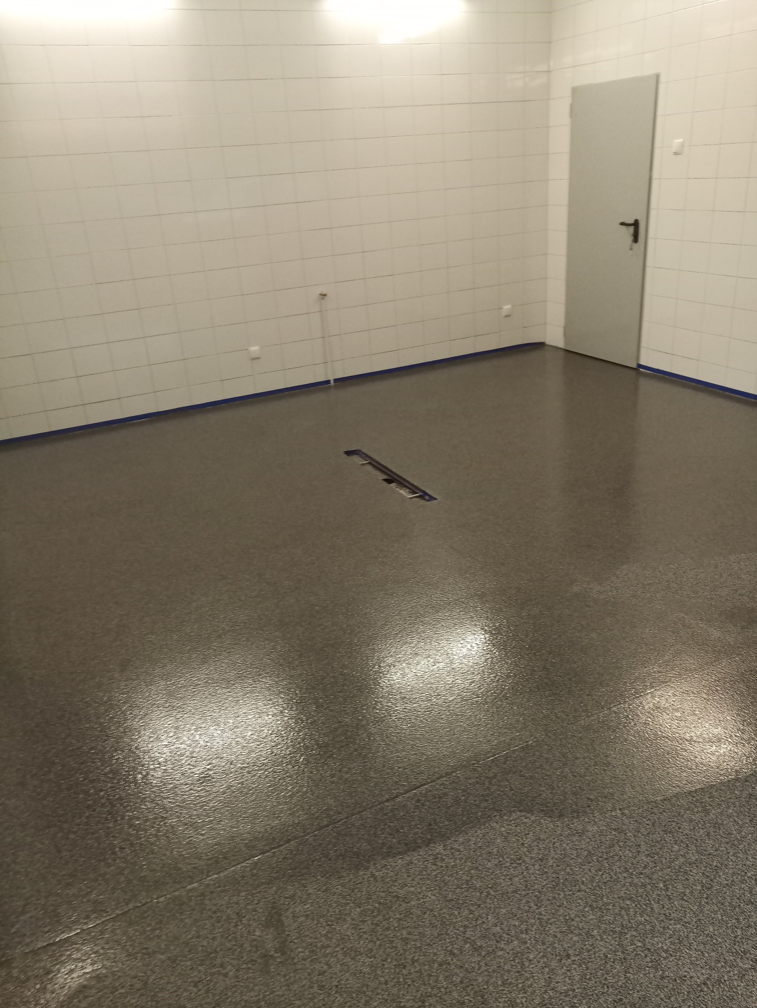 Szara, błyszcząca posadzka w pomieszczeniu wyłożonym białymi płytkami, widoczny odpływ liniowy i niebieska taśma malarska przy ścianach.