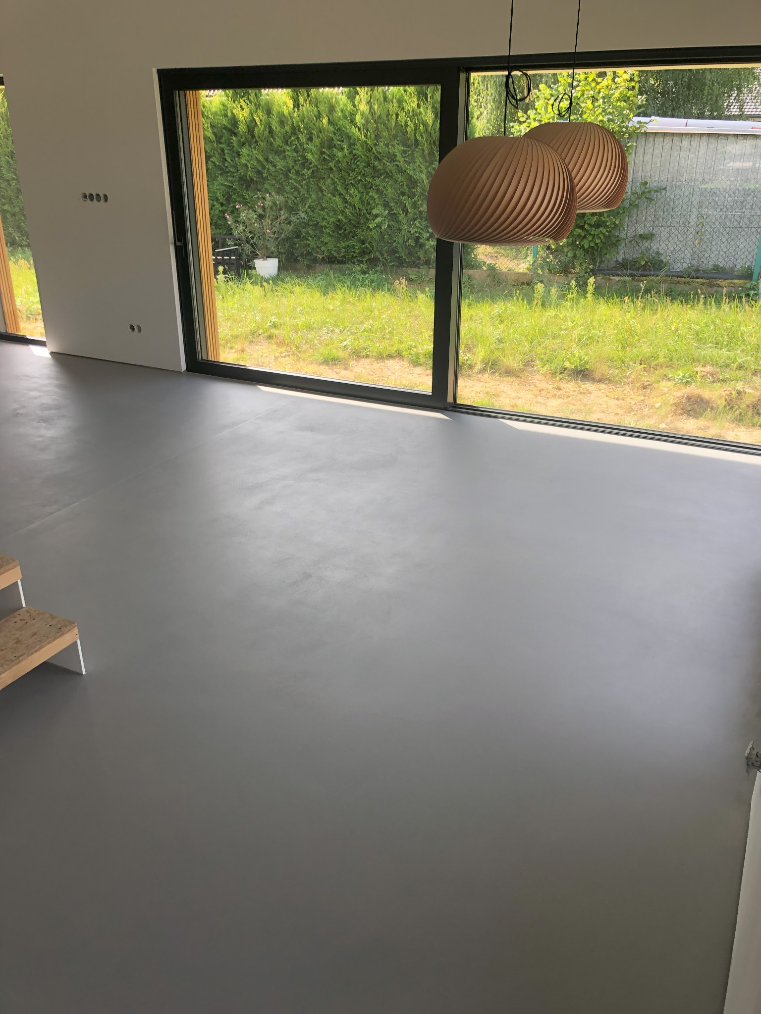 Szara, gładka posadzka w nowoczesnym wnętrzu z dużym oknem na ogród. Dwa dekoracyjne, drewniane abażury wiszą nad podłogą. Minimalistyczny design.