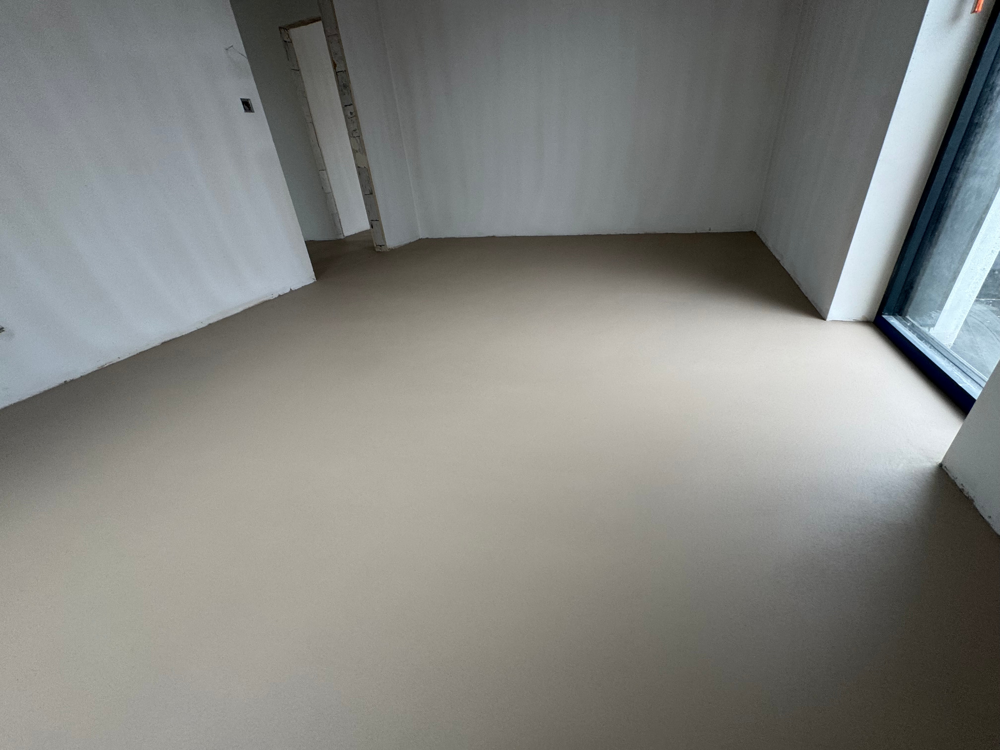 Jasna, gładka posadzka w surowym wnętrzu z oknem. Minimalistyczny design, neutralna kolorystyka, widoczne niedoskonałości ścian. Ukończona realizacja.