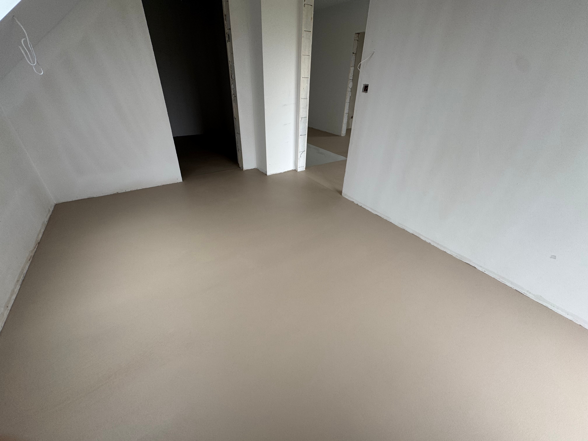 Beżowa, gładka posadzka w surowym wnętrzu z białymi ścianami i otworami drzwiowymi. Minimalistyczny design, neutralna kolorystyka, widok na sąsiednie pomieszczenia.