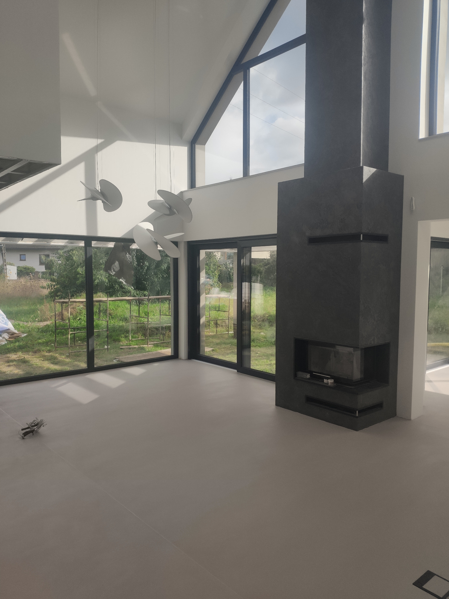 Jasna, minimalistyczna przestrzeń z szarą posadzką i kominkiem obłożonym ciemnym kamieniem. Duże okna z widokiem na zieleń. Nowoczesne lampy wiszące.